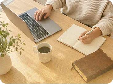 Person writing in a notebook beside a laptop on a wooden desk with a Bible